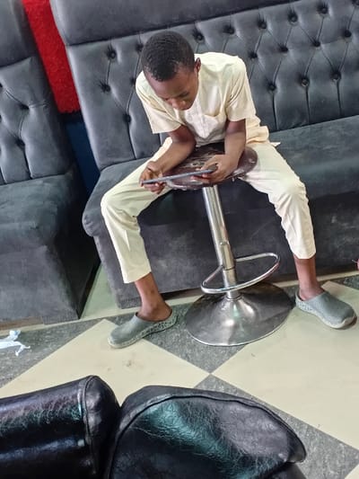 A young male is seated on a round, brown-cushioned bar stool with a chrome-finished pole and base, looking down at and operating a dark-colored smartphone held horizontally in both hands. He is dressed in a light-beige short-sleeved tunic with a chest pocket and matching trousers that end above the ankles, paired with grey perforated slip-on shoes. Behind him is a dark grey tufted sofa featuring high, button-backed sections, with a vertical red surface visible immediately to its left. In the lower foreground, the tops of two black upholstered sections, likely part of another sofa or chairs, are partially visible. The floor is covered with a checkerboard pattern of light cream and grey tiles. A small, crumpled white object lies on the floor to the left. The scene is an indoor setting in Dorayi, Nigeria.