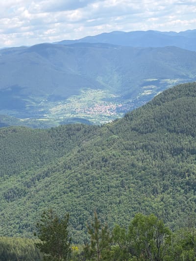 This is a high-angle, long-shot, natural landscape photograph taken on a cloudy day. The scene depicts a vast expanse of rolling, tree-covered mountains, with a village nestled in a valley in the middle ground. The foreground features dense, dark green evergreen trees on a steep slope. In the middle ground, the valley opens up, revealing a cluster of buildings with red-tiled roofs that form a village. Beyond the village, more mountains are visible, fading into a hazy blue in the distance. The sky is filled with white, fluffy clouds against a light blue background. The overall impression is one of a serene, rural mountainous region. The location is identified as Kosovo, Bulgaria.