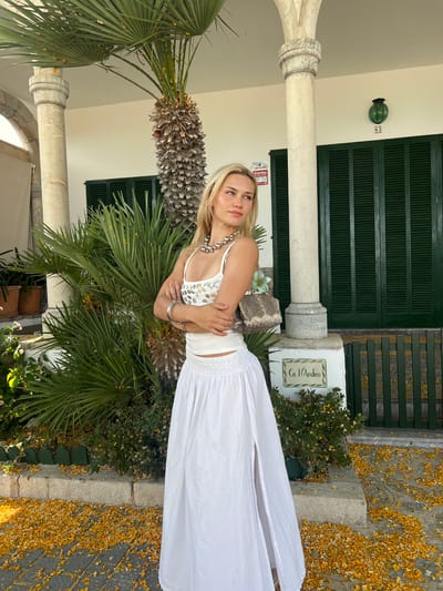 A blonde woman stands with her arms crossed in front of a building in Port De Pollença, Spain. She is wearing a white crop top adorned with iridescent sequins and a long, flowing white skirt with a thigh-high slit. She accessorizes with a large, chunky silver necklace and bangles on her wrists. A small, textured grey and beige handbag hangs from her left shoulder.

The background features the facade of a building with large, dark green louvered shutters and white columns. A tall palm tree and lush green plants surround the entrance. The ground is paved with stone and is heavily scattered with fallen yellow petals. A sign on the building reads "Ca N'Andreu" and another small sign displays the number "83". A security camera is visible mounted on the wall. The overall impression is one of a sunny, Mediterranean setting.