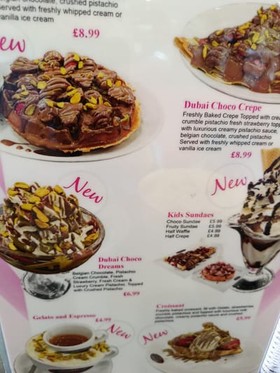 This is a close-up, eye-level, interior shot of a menu for a dessert shop. The menu features a variety of sweet treats with colorful images and descriptions. The focus is on various chocolate and pistachio-based desserts, including crepes, sundaes, waffles, and a gelato and espresso combination. Some items are marked as "New." Prices are listed in pounds (£). The background is a clean, light-colored surface, likely a table or counter. The lighting is bright, suggesting an indoor setting during the day. The overall tone is enticing and aims to showcase the appealing nature of the desserts. The menu items include "Dubai Choco Crepe," "Kids Sundaes," "Dubai Choco Dreams," "Gelato and Espresso," and "Croissant." Specific ingredients mentioned are Belgian chocolate, pistachio, strawberries, cream, and vanilla ice cream.