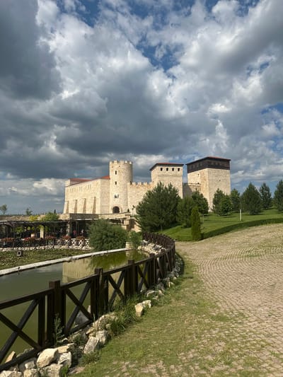 The image depicts a medieval-style castle with stone walls and turrets, situated in a landscape with a pond, manicured lawns, trees, and a paved walkway. The sky is partly cloudy. Several people are visible in the distance, appearing to be near a covered seating area by the pond. The architecture of the castle suggests it might be a tourist attraction or a historical replica. The location is identified as Neofit Rilski, Bulgaria.