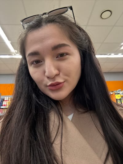 A close-up, front-facing photograph captures an adult female in an indoor retail setting within the city of Kurovskoye, Russia. The subject has long, dark brown hair with black-framed sunglasses positioned on top of her head. Her facial features include dark eyebrows, dark eyes, and lips colored with a reddish-brown matte lipstick, slightly pursed. She is wearing a light brown, possibly camel-colored, coat over a white undergarment. The background features a white tiled ceiling with visible fluorescent strip lights and at least one circular recessed light fixture. To the left, shelves stocked with various bottled or canned products are visible. To the right, an orange display or wall with a yellow price tag showing "79" is discernible.
