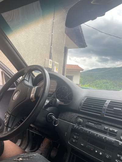 The image is an interior view of a car, likely a BMW, looking out through the windshield. The dashboard and steering wheel are visible, with a key in the ignition. Outside the car, a building with a red-tiled roof is seen, along with a hillside covered in green trees under a cloudy sky. The overall impression is of a rural or suburban setting. The location is identified as Vlahovo, Bulgaria.