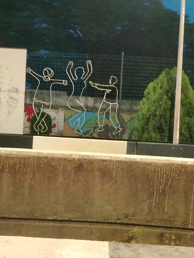 The image depicts a nighttime scene in Ikoyi, Nigeria, featuring a mural on a wall. The mural is outlined in white and depicts three stylized figures in dynamic poses, appearing to be dancing or celebrating. The figures are abstract, with simple lines representing their bodies and limbs. Behind the figures, a mosaic pattern is visible, composed of colorful geometric shapes. A black metal fence runs horizontally across the background, separating the mural from a dark, leafy tree on the right. In the foreground, a concrete barrier runs along the bottom of the frame, with a lighter-colored section on the very bottom. The overall impression is of public art displayed in an outdoor setting at dusk or night.