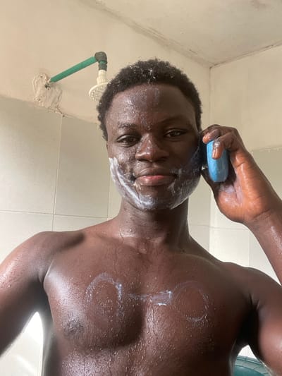 A young adult male, shirtless and wet, is shown taking a selfie in a bathroom located in the city of Mosafejaw, Nigeria. He has dark, short, curly hair and is smiling directly at the camera. White lather is present on his face, specifically on his cheeks and chin, and two distinct circular patterns of lather are visible on his wet chest. His left hand holds a blue bar of soap to his left ear. In the background, a white showerhead is mounted on a light-colored wall via a green pipe, with the lower wall section tiled in a lighter shade. The ceiling above shows signs of discoloration.