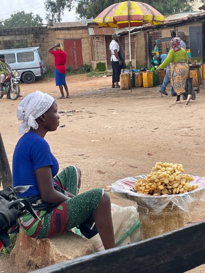 In Wang, Nigeria, a woman with dark skin, wearing a vibrant blue t-shirt and a patterned green and red skirt, sits on a stone in a dusty outdoor market. She has a white headscarf tied around her head. In front of her, on a raised platform covered with plastic sheeting, is a large pile of small, circular, fried food items. To her left, a black motorcycle is partially visible. In the background, other market activities are taking place. A person in a red shirt and blue skirt stands near a building with red doors. Further in the background, several yellow and brown jerry cans are stacked, and a small, three-wheeled vehicle is visible. Another woman, wearing a green top and a patterned skirt, stands near an umbrella-covered stall. The ground is dirt and sand, with scattered debris. The sky is overcast.