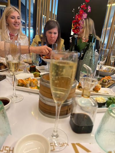 A brightly lit restaurant scene shows three women seated at a table with sushi, drinks, and decorative flowers, most likely in Torreforta, Spain, based on visual cues. Two women are in the foreground: one with blonde hair, wearing a white top with a red necklace, and smiling; the other, with brown hair and a yellow top, is holding food in her hand, appearing to eat. A third woman with blonde hair is visible further back, behind flowers and various glass bottles. The table is filled with sushi plates, champagne glasses, soy sauce containers, and wooden barrel-shaped objects. The background shows vertical metal bars and a white patterned wall.