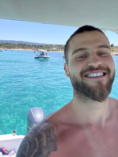 In this medium shot, a man with short dark hair and a beard smiles at the camera from a boat. He is shirtless, and the sunlight highlights his tanned skin. The water around the boat is clear and turquoise, with sunlight reflecting off its surface. In the background, other boats are visible, as well as a hilly coastline with green vegetation and some white buildings. The sky is a clear blue. The scene appears to be in a Mediterranean coastal area, likely Greece, given the clear blue water and the architecture visible on the shore.