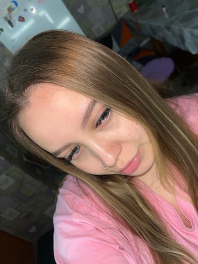 A close-up, slightly rotated image shows a light-skinned person with light brown hair and a silver nose ring. The person is wearing a pink shirt. The background includes a table surface with stickers and other objects, and a patterned surface. The image provides no information about the city of Port-Katon, Russia.
