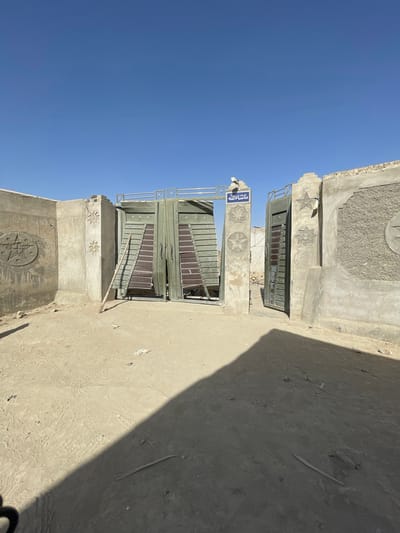 This image presents an exterior view of an entranceway in Spin Boldak, Afghanistan, under a clear, cloudless blue sky. The foreground consists of an uneven, light brown sandy or dirt ground with faint tracks and scattered small debris, including thin wooden sticks. A concrete wall runs along the left side, extending to form an entrance structure.
The central feature is a partially damaged entranceway comprising two robust concrete pillars supporting double-leaf metal gates, painted a faded olive green. The gates are slightly ajar, with their internal slat-like construction partially visible. A light-colored wooden prop leans against the left gate leaf. A simple metal railing connects the tops of the two gate pillars. Above the left gate pillar, a blue rectangular sign with white Arabic script reads "ู
ุงุดุงุกุงููู" (Mashallah).
Both the concrete walls and pillars are rough-textured and unpainted, appearing weathered. The wall sections and pillars feature embedded decorative elements: circular plaques containing raised five-pointed stars (pentagrams). The right wall segment also includes a textured, pebbled-dash-like finish on its upper portion. Strong shadows from the left-side wall and foreground objects are cast onto the ground, indicating bright overhead sunlight. Beyond the gates, a desolate, dusty landscape with distant, undefined structures is faintly visible. No individuals or active subjects are present in the scene.