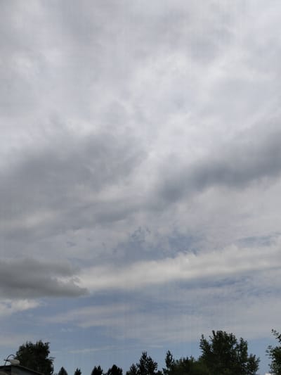 The image shows a cloudy sky above a treeline in Bulaev, Kazakhstan. The sky is filled with various shades of gray clouds, indicating an overcast or partly cloudy day. In the lower portion of the image, the dark green tops of coniferous and deciduous trees are visible, forming a natural border against the sky. A small portion of a building's roof and a street lamp are also visible on the left side of the frame, suggesting an urban or suburban setting within Bulaev. There are no people or active interactions depicted in the scene.