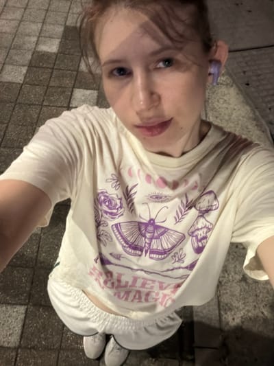 This is a selfie taken outdoors at night, possibly in a city like Dongducheon-si, South Korea. The subject is a young person with light skin and blonde hair, looking up at the camera. They are wearing a cream-colored t-shirt with a purple graphic print featuring a moth, roses, an eye, and some text. The t-shirt also has moon phases printed on it in pink. The person is also wearing white shorts and light-colored sneakers. The ground is paved with rectangular tiles, and there are shadows suggesting artificial lighting. The overall mood is casual and perhaps a bit playful, with the subject's mouth slightly open. The visible text on the t-shirt appears to read "Believe in Magic."