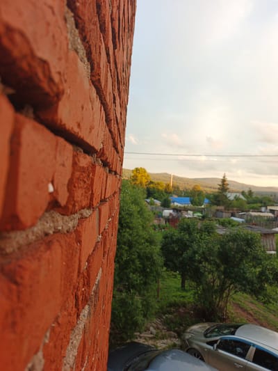 A close-up of a red brick wall in the foreground dominates the left side of the frame, with the texture of the bricks and mortar clearly visible. Beyond the wall, the scene opens up to a rural village setting in Medvedyovka, Russia. The background depicts a collection of houses, some with blue roofs, nestled amongst lush green trees. Rolling hills and mountains form the backdrop, suggesting a natural, somewhat remote location. The sky is overcast with soft clouds, indicating it is likely late afternoon or early evening, with the light casting a gentle, diffused glow on the landscape. A couple of cars are parked on a dirt or gravel area in the middle ground. There are no people visible in the image, and no text is present. The overall impression is one of a quiet, rural evening in a small Russian village.