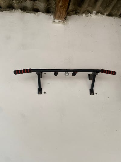This media file shows a black pull-up bar mounted horizontally on a white, cracked plaster wall. The pull-up bar has red and black padded grips at either end and also in the middle. A silver metal ring is attached to the underside of the bar, likely for attaching other exercise equipment. The wall appears to be in an unfinished or rustic setting, with exposed wooden beams and corrugated roofing visible on the left side of the frame. The lighting suggests it might be daytime, but there are no clear indicators of time of day or weather. The overall impression is of a functional workout station in a somewhat dilapidated or utilitarian space. No visible text is present in the image.