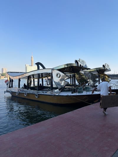 The image depicts a boat shaped like a fish, docked alongside a red-colored walkway. The boat features a gleaming, metallic exterior with multiple fish-shaped decorative elements. One person, wearing a white t-shirt, is standing on the boat, seemingly engaged in some activity. The boat is on the water, with buildings visible in the background, including a distinctive tower. The sky is clear and blue, suggesting a bright, sunny day. The overall scene appears to be a waterfront location, likely a tourist attraction or a place for boat tours. The boat itself is a prominent object, designed to be eye-catching. The walkway has a textured surface, resembling wood grain. The image is taken from a slightly elevated angle, looking down at the boat and the water. The presence of buildings and the clear sky suggest this is likely daytime. There is some text visible on the boat's side, but it is not legible in this image. The boat's design suggests a potentially festive or whimsical atmosphere.