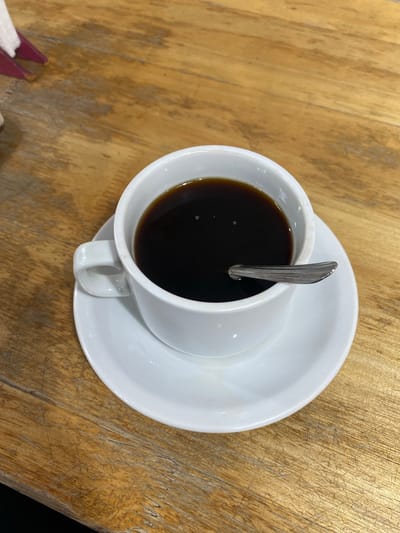 The media file presents an overhead view of a cup of black coffee, resting on a white ceramic saucer. A silver spoon is placed vertically within the dark liquid of the coffee. The cup and saucer are clean and white, providing a stark contrast to the deep, dark brown color of the coffee.
These items are situated on a wooden table, which appears to be light brown or amber in color. The table surface is notably rustic, featuring distinct wood grain patterns and numerous visible scratches and marks, indicating wear and tear. A small, blurred, purple object, possibly a napkin holder or a folded menu, is partially visible in the bottom-left corner of the frame.
The lighting is even, suggesting an indoor setting without harsh shadows. No individuals are depicted in the image. The implied activity is the imminent or ongoing consumption of coffee. No text is visible within the frame. The context for this scene is Monte Grande, Argentina.
