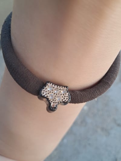 An extreme close-up shot shows a dark gray fabric hair tie encircling a person's arm. Attached to the hair tie is a small, black, star-shaped charm adorned with numerous clear rhinestones. The skin tone of the arm suggests it belongs to a person with fair to medium complexion. The background is out of focus, showing hints of a textured surface, possibly concrete or stone, and a light gray, possibly blurred background. The lighting appears to be natural and somewhat diffused, creating soft shadows on the arm.