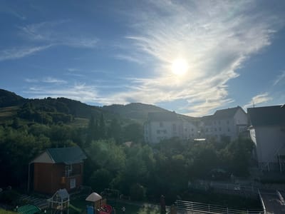 A bright, sunlit day in a rural area, possibly a resort or village, with a focus on a cluster of buildings and a playground set against a backdrop of rolling, tree-covered hills. The sun is high and slightly to the right of the frame, creating a bright glare and casting a warm light across the scene. Wispy white clouds are scattered across the clear blue sky.
In the foreground, a small, two-story wooden house with a green roof is visible on the left. Adjacent to it is a children's playground with colorful equipment including a slide, swings, and play structures.
Further back, several larger multi-story buildings, likely hotels or apartment complexes, are nestled among lush green trees. These buildings are white or light-colored with visible windows and balconies. One building on the far right has an external staircase.
The landscape is characterized by dense, green vegetation covering the hills. A road or path is visible leading through the area, and a white railing lines a pathway in the lower right quadrant of the image. A few indistinct figures, possibly people, can be seen in the distance, near the buildings and on the path. The overall impression is of a tranquil, scenic location.