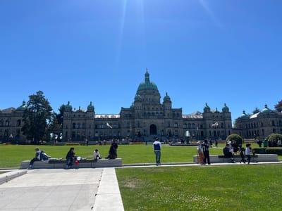 A wide shot captures the majestic facade of the British Columbia Parliament Buildings in Victoria, Canada, on a bright, sunny day. The imposing stone structure, adorned with green domes and intricate architectural details, dominates the background under a clear blue sky. Sunlight glints off the building and the sky, indicating mid-day.
In the foreground and middle ground, a lush green lawn is dotted with small white clover flowers. Several groups of people are scattered across the lawn and sitting on low, light-colored stone benches. On the far left, a group of three people are casually seated, with one person lying down, suggesting a relaxed atmosphere. Closer to the center, a family sits together, with a child held by an adult, and a red and white balloon visible. Other individuals and small groups are also seen enjoying the outdoors, some standing, others seated.
Towards the center of the image, a large fountain sprays water, adding to the lively scene. Flags fly from poles in front of the Parliament Buildings. The overall mood is one of leisure and enjoyment of a beautiful day in a public space. The architecture, the setting, and the presence of visitors suggest this is a popular tourist destination. No visible text is present in the image.