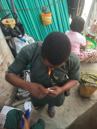 The image depicts an outdoor or semi-outdoor scene in Ajah, Nigeria, likely a market stall or roadside area. In the foreground, a dark-skinned male, viewed from above, wears a dark green tunic and dark blue flip-flops. He has short, curly dark hair and is focused on manipulating a small white cylindrical object, possibly a cigarette labeled "Italian," with both hands. A small screwdriver with a yellow and red handle is clipped to his shirt. A black shoulder bag strap is visible across his chest. To his lower left, a person's legs, clad in dark green trousers, are partially visible, holding a blue-cased smartphone.
In the mid-ground to the right, a dark-skinned female, seen from behind, is seated on a low stool. She wears a light pink t-shirt and patterned trousers, and her hair is styled in dark braids. She is actively sorting light-colored items, likely peanuts, in a green plastic bowl placed in her lap. Next to her, a small wooden stool holds a silver circular tray with several red-capped bottles containing liquid, presumably beverages. A yellow bucket, partially covered with a patterned cloth, rests on the ground near her.
The background features a vertical corrugated metal wall or fence, painted or covered in a bright green color. Hanging from a chain attached to this wall is an orange plastic bucket with "Goldberg Custard Powder" clearly visible on its side. To the left, against a light-colored wall or partition, are several black and white plastic bags and some debris. The ground throughout the scene is a mix of concrete and packed earth.