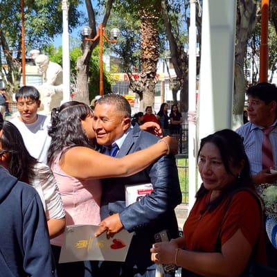A family is celebrating an event in a park in Cerro Colorado, Peru. A man in a suit holds up a certificate that says "Municipalidad Distrital Cerro Colorado." He is being kissed by a woman. In the background, other families are present, and trees and buildings can be seen.