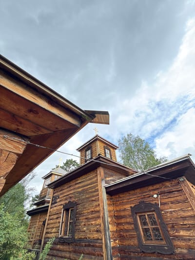 The image shows an exterior view of wooden buildings with Russian Orthodox crosses on their roofs. The buildings are constructed from logs, with visible wood grain and some signs of weathering. One of the structures features a prominent window with an ornate wooden frame. The scene is set against a cloudy sky and surrounded by greenery, suggesting an outdoor location. Based on the architectural style and religious iconography, this is likely a church or monastery in a rural or semi-rural setting. Without specific visual cues to pinpoint the exact location within Chemal, Russia, further details regarding the city cannot be provided. No people or discernible actions are present in the image.