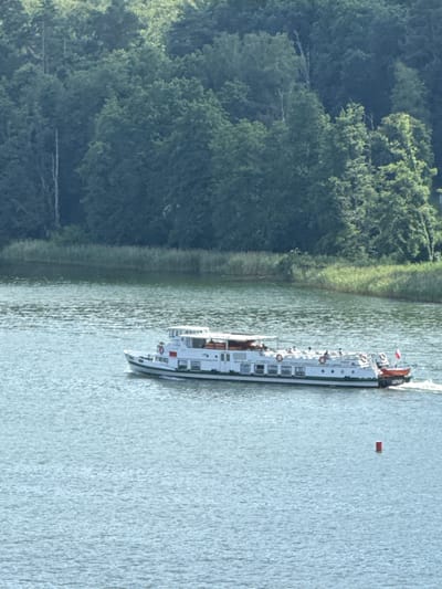 The image shows a white passenger boat with several decks, sailing on a body of water. The boat has multiple windows and life preservers visible on its side. A small red and white flag is flying at the stern of the boat, along with what appears to be the number "G02/420" printed on the back. The boat is moving, creating ripples in the water. In the distance, a small red buoy is visible. The left side of the image features a dense green forest with tall trees, suggesting a natural, possibly rural, setting. The water is calm with some texture from the boat's movement and possibly a gentle breeze. The lighting suggests it is daytime, with sunlight illuminating the scene. The overall atmosphere is peaceful and scenic, characteristic of a leisurely boat trip on a lake or river. The location context provided, Mikołajki, Poland, indicates this scene is likely taking place in the Masurian Lake District, a popular tourist area known for its waterways and forests.