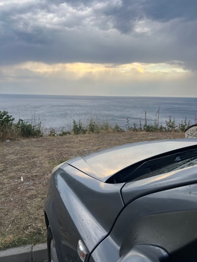 The image depicts a close-up view of a grey car's hood and fender, with the sea and a cloudy sky in the background. The car's surface is wet, with visible water droplets. The foreground shows dry, sparse vegetation and a curb. The sea is calm, and sunlight is breaking through the clouds, creating rays of light over the water. The scene appears to be an outdoor parking area overlooking the sea, possibly in a coastal town. The location is identified as Pečurice, Montenegro. No people are visible in the image.