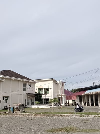 The image depicts an outdoor scene in Mesjid Reuntoh, Indonesia, under an overcast sky. In the foreground, a gravel-covered area stretches across the bottom of the frame. Beyond the gravel, a patch of dry grass and some sparse vegetation can be seen.
Dominating the middle ground are several buildings. On the left, a white building with a dark-pitched roof features a modern architectural style with multiple windows. To the right of this building, another white building has a green-trimmed facade and balconies. Further to the right, a long, open-sided structure with a reddish-brown roof and white pillars is visible, with vehicles parked inside. A smaller building with a maroon roof and white walls is situated between the white buildings and the open-sided structure.
Several people are present, mostly on motorcycles. On the far left, two individuals are seated on motorcycles parked near the first white building. In the middle of the scene, a person is riding a black motorcycle, wearing a white shirt and black pants, and a hijab. Other motorcycles and individuals are scattered throughout the area, some parked and others in motion. A small, open-sided shelter with a rusted roof is located in the middle ground. Overhead, power lines crisscross the sky. The overall impression is a functional, possibly institutional or commercial, area with ample parking space.