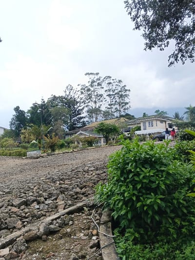 The image captures an outdoor scene featuring a rocky, unpaved path leading upwards, flanked by lush greenery and several buildings in a hilly or mountainous setting under an overcast sky.
In the immediate foreground, the ground is covered with a rough surface of various sizes of rocks, gravel, and dirt, suggesting an unmaintained road or pathway. This rocky terrain extends from the bottom of the frame, curving slightly upwards and to the left. Along the right edge of this path, a low concrete or stone border is visible, with some white wires running along it. Adjacent to this border, on the far right, a vibrant green bush with dense, leafy foliage fills a significant portion of the foreground, partially obscuring the path.
In the midground, several buildings are nestled among abundant trees and vegetation. To the right, a prominent two-story building with light-colored (possibly cream or white) walls and a dark roof features multiple windows, many with white frames, and a white veranda or balcony running along its front. A black car is parked near this building, and two figures, appearing to be women, are standing next to it; one wears a red top and the other a lighter-colored top. Further to the left, another building with a distinct white decorative trim along its roofline is visible. Various trees and bushes of different heights and shades of green are interspersed throughout the midground, including a tall lamppost or utility pole surrounded by trees on the left side. Some of the trees are quite tall and slender with foliage concentrated at their tops, indicative of certain species like eucalyptus.
The background is dominated by a largely overcast sky, appearing light greyish-white, indicating a cloudy day. Faintly visible on the distant horizon are rolling hills or mountains, covered in vegetation, blending into the cloudy atmosphere. The overall impression is one of a serene, possibly rural or semi-rural, residential area in an elevated or mountainous region, characterized by its natural greenery and rugged terrain. There is no discernible text visible in the image.