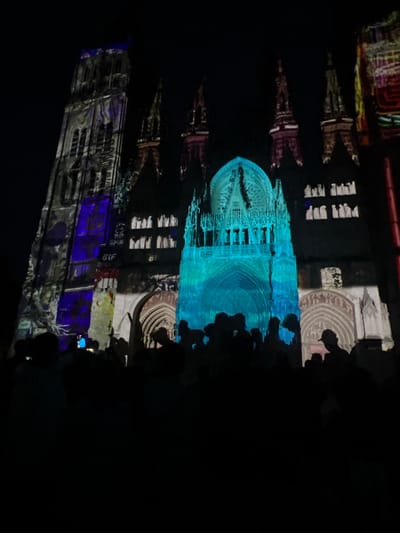 The image depicts the facade of Rouen Cathedral in Rouen, France, at night, illuminated by a vibrant light projection show. The grand Gothic architecture of the cathedral is visible, overlaid with dynamic colors and patterns. The tall left tower (Tour Saint-Romain) is predominantly lit with shades of purple and grey, displaying abstract designs and faint text including "61F" and "GIF". The central section of the cathedral, encompassing the main portal and rose window, glows intensely in various shades of blue and turquoise, highlighting its intricate carvings and arched structures. Towards the right, other spires and sections of the facade are illuminated with warmer hues of red, brown, and yellow, presenting distinct, colorful patterns.

In the foreground, a large crowd of spectators is visible in silhouette, positioned directly in front of and looking up at the illuminated cathedral. Their figures are almost entirely dark against the bright projections. Several individuals can be identified by their head and shoulder outlines, with at least two individuals on the left holding up illuminated smartphones, likely recording the event. One person on the far right is clearly identifiable by a baseball cap. The sky above the cathedral is uniformly dark, emphasizing the illuminated architectural display.