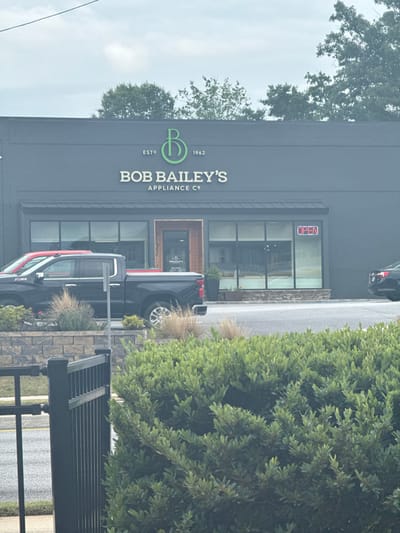 The image depicts the storefront of "Bob Bailey's Appliance Co." in Stone Mountain, USA. The building is a dark gray, one-story structure with a prominent logo and signage. An "OPEN" sign is visible in the window. Two dark-colored pickup trucks are parked in front of the store. Landscaping elements including bushes and decorative grasses are also visible. The weather appears overcast.