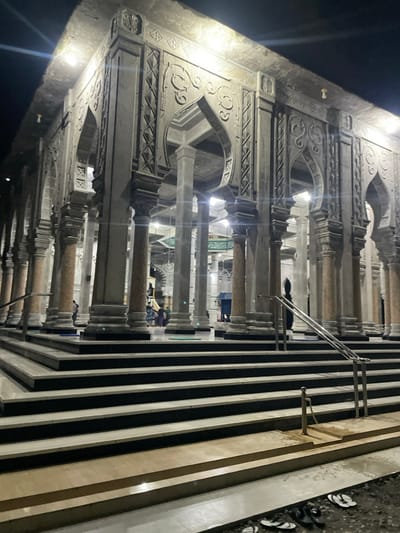 A building with intricate architectural details features rows of columns connected by arches. The stone structure is adorned with decorative carvings. Parallel black and white stripes run alongside the colonnade, possibly delineating a walkway or steps. Shoes are clustered on a surface next to the stripes. The area is illuminated, suggesting it might be nighttime. People are visible within the structure.