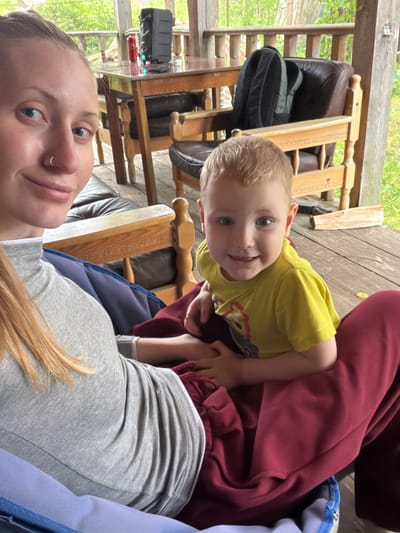 The image depicts a young woman and a small child seated on a porch. The woman, with blonde hair and a nose ring, is wearing a grey turtleneck. The child, with blonde hair and blue eyes, is wearing a yellow t-shirt. They are both covered by a dark red blanket. In the background, a wooden table and chairs, a backpack, and a speaker are visible. The environment suggests an outdoor setting, possibly a rustic cabin or a porch in a wooded area. There is no indication of the location being Merkuriy, Russia.