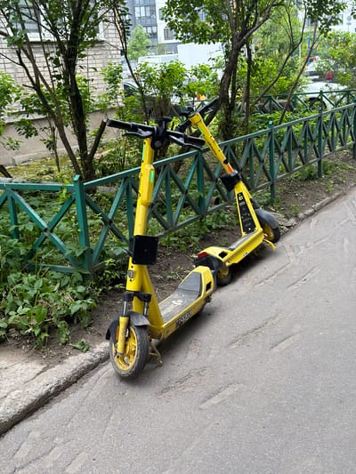 The image displays two bright yellow electric scooters positioned outdoors next to a green metal fence. Lush green trees and shrubs are visible behind and around the fence, with residential or urban buildings partially discernible through the foliage in the background. An asphalt path or road runs parallel to the fence. One scooter leans against the fence, while the other lies on its side, partially resting on the ground and against the fence, suggesting they are parked or discarded.
The scene is set during the daytime, under what appears to be an overcast or cloudy sky, indicated by the diffused lighting and lack of strong shadows. The scooters show signs of regular use, with some dirt on their wheels and lower parts. The asphalt path features some irregular indentations.
Prominently displayed on the side of both scooters is the brand name "Яндекс Go" (Yandex Go), identifying them as part of a shared electric scooter service. A partial alphanumeric code, "B53K", is discernible on the rear of one scooter. No people are present in the image. The setting appears to be an urban or suburban area in Novosergievka, Russia.