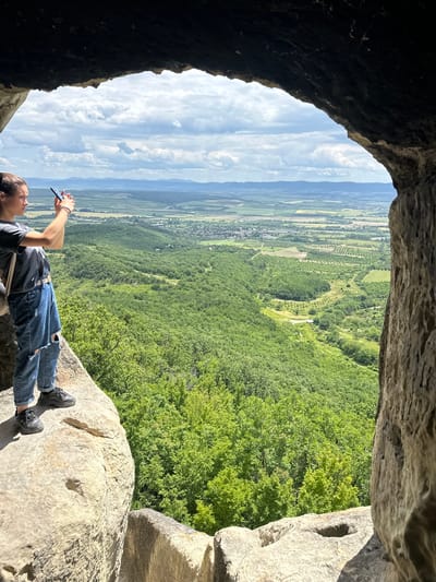 A person lies on a rock ledge, oriented horizontally in the frame, looking out from a cave towards a broad, panoramic view of fields, forests, and distant mountains under a cloudy sky. The person wears blue jeans and a dark shirt, and is holding a cell phone, seemingly taking a picture. The rocky opening of the cave frames the scene, with rough, dark rock on the left and lighter, textured rock on the right. The bright outdoor light contrasts with the interior darkness of the cave. The location is Han Krum, Bulgaria, as revealed by the GPS coordinates embedded in the image file.
