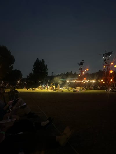 This image depicts an outdoor night event on a large grassy field in Burbank, California, United States. The scene is dominated by a distant, brightly illuminated stage on the right, where musical instruments, including a drum set, are visible under yellow-white stage lights. A faint haze or smoke rises from the stage area.
In the midground, two tall, temporary truss structures serve as lighting towers, each equipped with multiple spotlights and several large, spherical, orange-glowing lanterns. Orange string lights with similar lanterns extend horizontally from these towers across the event area. Numerous spectators are scattered across the dark grassy field, primarily seated, observing the stage.
In the immediate foreground, to the lower left, two individuals are partially visible, seated in dark, foldable chairs. One person, wearing light-colored pants and dark footwear, holds a white disposable bowl and utensil, indicating consumption of food. The legs and light-colored footwear of a second individual are visible to the right of the first.
The background consists of dark, tree-covered hills and a dark, cloudless night sky. Tall trees are silhouetted against the sky on the left and center. The overall environment is an open-air venue, likely a park or event space, hosting a live performance under artificial illumination.