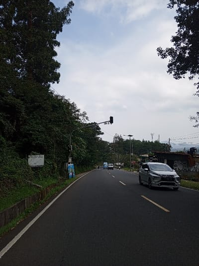 The image displays a two-lane asphalt road in Ciloto, Indonesia, under an overcast sky. The road, appearing damp, curves slightly left and is flanked by dense vegetation. On the immediate left, a white solid line marks the road edge, followed by a low concrete barrier. Beyond this, an embankment is covered in dark green foliage, partially obscuring a white rectangular sign with dark text and a graphic. A utility pole on the left supports an inactive black traffic light suspended over the road, with a smaller blue-and-white utility box attached to the pole below.
The road features a solid yellow center line and a broken yellow line further right. A silver Mitsubishi Xpander Multi-Purpose Vehicle (MPV) is in motion on the right side of the road, traveling away from the viewer. In the opposing lane, further down the road, a light blue truck and a smaller white vehicle are visible. A motorcycle is partially obscured by the utility pole on the left shoulder.
On the right side of the road, a dilapidated structure with corrugated metal components and white and black graffiti on its lower walls stands adjacent to the road. Beyond this structure and in the background, a street light with a solar panel, communication towers on a distant ridge, and a body of water (likely a lake or reservoir) are visible beneath the cloudy horizon. No identifiable individuals are present.