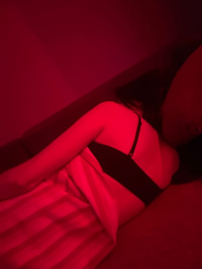 A person, possibly female, is lying on a couch, facing away from the camera and partially covered by a striped blanket. The scene is illuminated by red light, casting a strong red hue over the entire image and obscuring many details. The person is wearing a black bra. The background appears to be a wall, also under the red light. No specific location in Gliwice, Poland can be identified from the image.