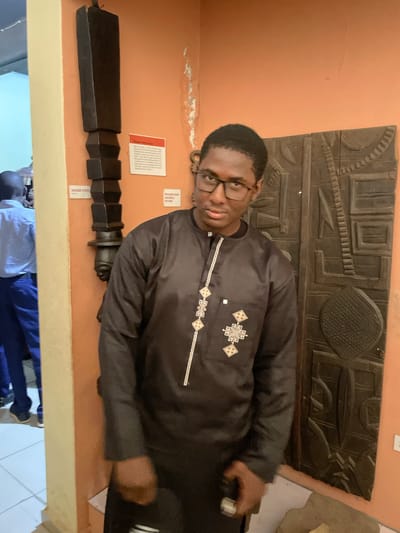 In Unguwar Shanu, Nigeria, a young Black male wearing glasses and a black traditional tunic stands in front of an orange wall. He is positioned slightly to the right of the frame. To his left, a tall, dark, carved wooden object, likely a house post, is mounted on the wall. Behind him, to his right, is a large, intricately carved wooden panel, also mounted on the wall. The wooden panel features geometric patterns and possibly figural elements. In the background, to the left of the house post, another person is visible, wearing a blue plaid shirt and blue trousers, facing away from the camera. The floor appears to be tiled. The overall scene suggests a museum or cultural exhibition space.