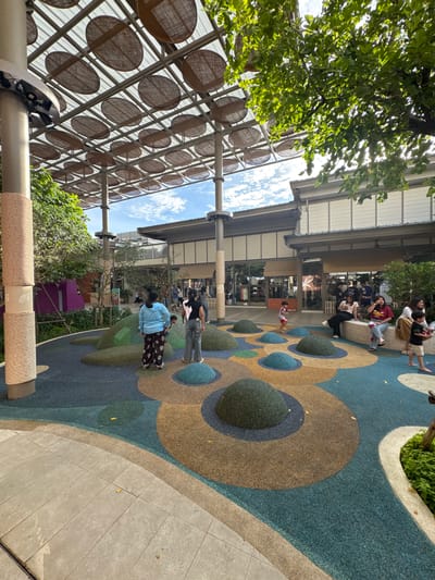 This media file depicts an outdoor scene in Krabi, Thailand, likely during the daytime given the bright sunlight filtering through the trees and reflecting off the structures. The weather appears pleasant and clear, with visible clouds in the sky.
The scene is a public space with a playground area featuring a colorful, patterned rubberized surface with circular and oval-shaped insets. Some of these insets are raised and covered in green, turf-like material, resembling small mounds. Adjacent to the playground is a tiled walkway. In the background, there are buildings with modern architecture, characterized by white facades and large windows. A prominent overhead structure is visible, composed of a metal frame supporting numerous circular, woven elements, possibly a decorative awning or ceiling. Lush green foliage and trees are scattered throughout the scene, indicating a well-maintained outdoor environment.
Several people are present in the image. In the foreground, a person wearing a blue shirt and patterned pants is standing. Further in the background, a group of people are seated on a low wall or bench, some interacting and others looking around. A child can be seen in the playground area. The overall atmosphere seems relaxed and leisurely, suggesting a public gathering or a casual outing. No discernible text is visible in the image.