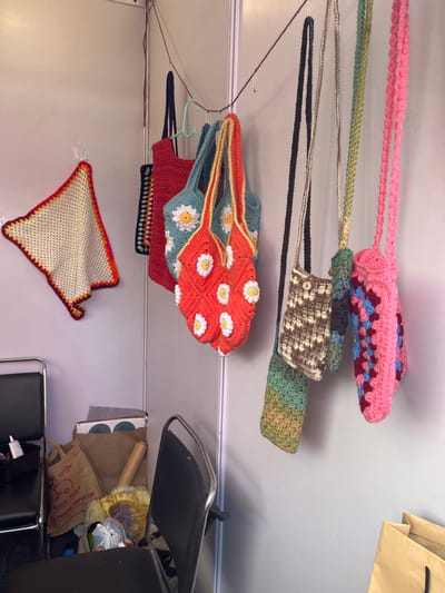 The image shows a variety of crocheted bags hanging on a string from a wall. The bags are all different colors and styles, and some have daisy-shaped designs. The bags are hanging on a wall in a room that contains a chair, a paper bag, and another crocheted piece. The location based on coordinates (latitude: 30.764, longitude: 76.798) is in Sector 17, Chandigarh, India.