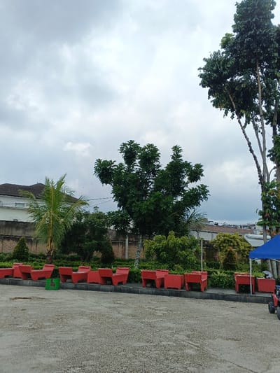 The scene depicts an outdoor paved area adjacent to a landscaped garden, located in Thành phố Biên Hòa, Vietnam, under an overcast sky. The foreground features a light-colored, textured concrete or asphalt surface. A row of twelve red, rectangular concrete planters or benches is arranged along a low curb, separating the paved area from a green garden bed. One green plastic bucket rests on the paved surface near the left end of the red structures.
The garden bed contains dense green foliage, including a small palm tree with bright green fronds on the left and a larger, mature tree with dark green leaves centrally positioned. Behind the garden, a light-colored concrete wall spans horizontally. To the far left, beyond this wall, a portion of a white building with a dark, sloped roof is visible. Taller trees with more sparse foliage are present on the right side of the garden. On the far right, a blue canopy is partially visible, under which a red and black children's ride-on toy vehicle and a white object (possibly a helmet) are situated. The sky is entirely covered by a uniform layer of light grey clouds. No individuals or active interactions are observed.