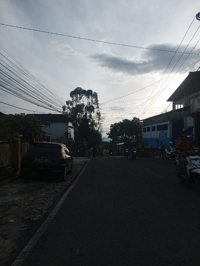 This image captures a street scene in Cipendawa, Indonesia, likely during the late afternoon or early evening under an overcast sky. The lighting is diffused, with the sun appearing low in the sky on the right, partially obscured by clouds.
The primary subject is a paved street, stretching from the foreground into the distance. On the left, a black car (license plate B 1537 JNB) is parked on a stone-paved shoulder next to a low, brown fence and dense green foliage. Behind the fence, a light-colored building with a dark roof is visible. Further down the street on the left, a tall, slender tree with prominent foliage stands out, surrounded by other trees and greenery.
On the right side of the street, a row of buildings lines the road. One building has a distinctive blue and white facade. The upper half of the image is dominated by a complex web of overhead utility wires crisscrossing the cloudy sky, emanating from unseen poles.
Several people are present, indicating ongoing activity. In the right foreground, a person wearing an orange shirt is seated on a white scooter. Other people, some on motorcycles and others on foot, can be seen further down the street, suggesting daily commuting or local movement. The scene depicts a typical residential or semi-commercial urban environment in Indonesia.