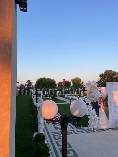 The image depicts an outdoor venue in Ratuș, Moldova, likely set up for a wedding reception. The setting sun casts a warm, orange glow on the left side of the frame, illuminating a portion of a building's exterior. In the foreground, two large, spherical light fixtures are visible, one casting a warm light. A dark metal lamp post stands between them.
The ground is paved with a geometric pattern of grey and white tiles. To the right of the paved walkway, a meticulously maintained green lawn is bordered by white stones and small, manicured evergreen bushes. Several white statues, resembling classical figures, are placed along the lawn's edge.
Further into the scene, a large, white banner is prominently displayed, featuring the text "WELCOME TO OUR WEDDING" and the date "30. 07. 2023." Below this, a seating chart is visible with names listed under table numbers. A large, white floral arrangement is positioned next to the banner, and a framed image of a bride and groom is integrated into the banner's design. In the background, lush green trees form a natural backdrop, and a red flag is visible on a pole in the distance. A clear, pale blue sky dominates the upper portion of the image.