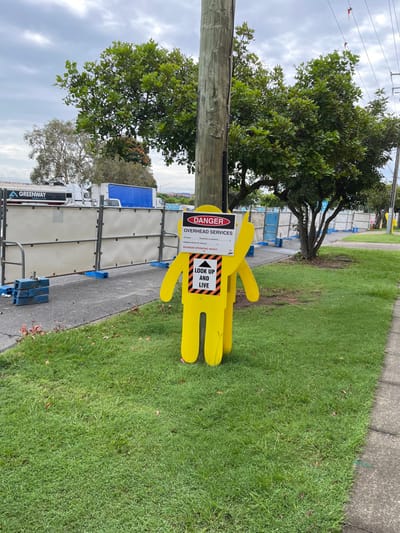 The image, taken in Hamilton, Australia, displays an outdoor scene featuring a prominent yellow, human-shaped cutout sign positioned on a strip of green grass adjacent to a paved sidewalk. The sign functions as a safety warning, with a red "DANGER" banner at the top and "OVERHEAD SERVICES" printed below it. Further down, there are sections detailing "MINIMUM HEIGHT OF POWERLINE CLEARANCE," "ISOLATION," and "MAXIMUM OPERATING HEIGHT," followed by numerical values, some of which are partially legible. A distinct black and yellow striped box on the sign prominently displays the message "LOOK UP AND LIVE" accompanied by an upward-pointing arrow.

Behind the sign stands a sturdy wooden utility pole, with overhead lines implied by the warning message. A large, leafy tree provides a verdant backdrop to the pole and sign. The sky is overcast, indicating a cloudy daytime setting. In the background, a series of temporary construction barriers or fencing lines what appears to be a work area. Behind these barriers, several vehicles are visible, including a truck with the word "GREENWAY" printed on its side. No people are present in the scene. The overall setting suggests a utility or construction work zone where a high level of caution is necessary due to the presence of overhead services.