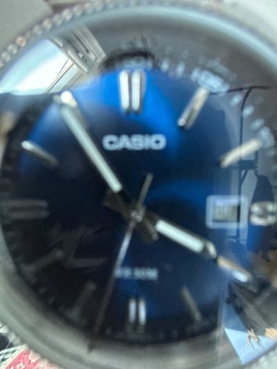 The image is a close-up, slightly blurry, macro photograph of a watch face. The watch has a dark blue dial and silver hands. The brand name "CASIO" is visible in white text on the dial. There are silver hour markers on the dial, some of which appear to be double-striped. A small digital display window is also visible near the top of the dial. The watch appears to be made of metal, with a textured bezel. The lighting suggests an indoor setting. Reflections are visible on the watch face, indicating a light source from above and to the left, possibly a window or lamp. The background is out of focus and appears to be a patterned surface, possibly a tablecloth or rug. There are no people or animals in the frame. The image is focused on the watch itself, highlighting its details. The overall impression is that of a detailed examination of a functional timepiece.
