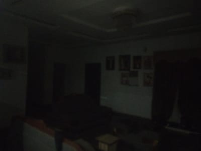 The image depicts a dimly lit interior scene, likely a living room or common area, within a building in Iyafin, Nigeria. The lighting is very low, suggesting it may be nighttime or the electricity is off. The room appears to be furnished with dark-colored furniture that is mostly obscured by shadow. On the wall in the background, several framed pictures or photographs are visible, arranged in a grid-like pattern. A ceiling light fixture is faintly illuminated. The floor is not clearly visible due to the poor lighting and shadows. The overall impression is one of darkness and a lack of clear detail.