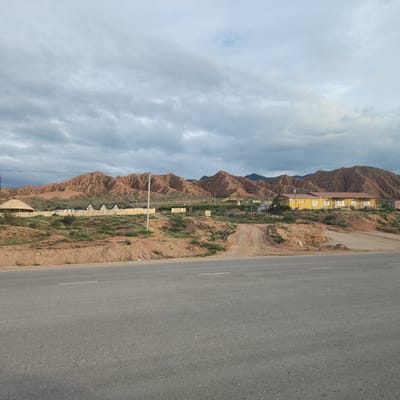 The image depicts a rural scene in Kaji-Say, Kyrgyzstan, under a cloudy sky. In the foreground, a two-lane asphalt road with a white dashed line runs horizontally. Beyond the road, a dry, reddish-brown landscape rises, dotted with sparse green vegetation.

On the rising terrain, a row of small, triangular-roofed structures, possibly cabins or glamping units, are visible behind a wooden fence. To the left of these structures is a conical-shaped building with a thatched roof. Further to the right, a long, low yellow building with a reddish-brown roof stands on a hill. The background is dominated by a range of rugged, eroded mountains with a distinctly reddish hue, characteristic of the region. The sky is filled with heavy, grey clouds, suggesting overcast or potentially stormy weather. The overall impression is one of a remote, developing tourist area situated in a dramatic natural landscape. There is no visible text in the image.