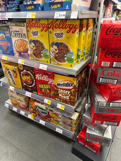 The image shows a supermarket shelf in Rotherham, United Kingdom, stocked with various breakfast cereals and drinks. On the top shelf, multiple packs of Coca-Cola Zero Sugar cans are stacked. Below, shelves are filled with breakfast cereals, including Kellogg's Coco Pops, Weetabix, Kellogg's Krave, Alpen, and Quaker Oats. Price labels are visible on the shelf edges, indicating the cost of each product. Other products on the bottom shelf include Squares and Shreddies.